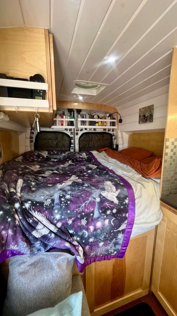 Inside of a converted cargo van with a fixed bed, overhead compartments, and bookshelves.