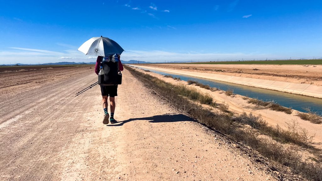 Backpacker walking a canal with an umbrella for shade on the Desert Winter Thru-Hike