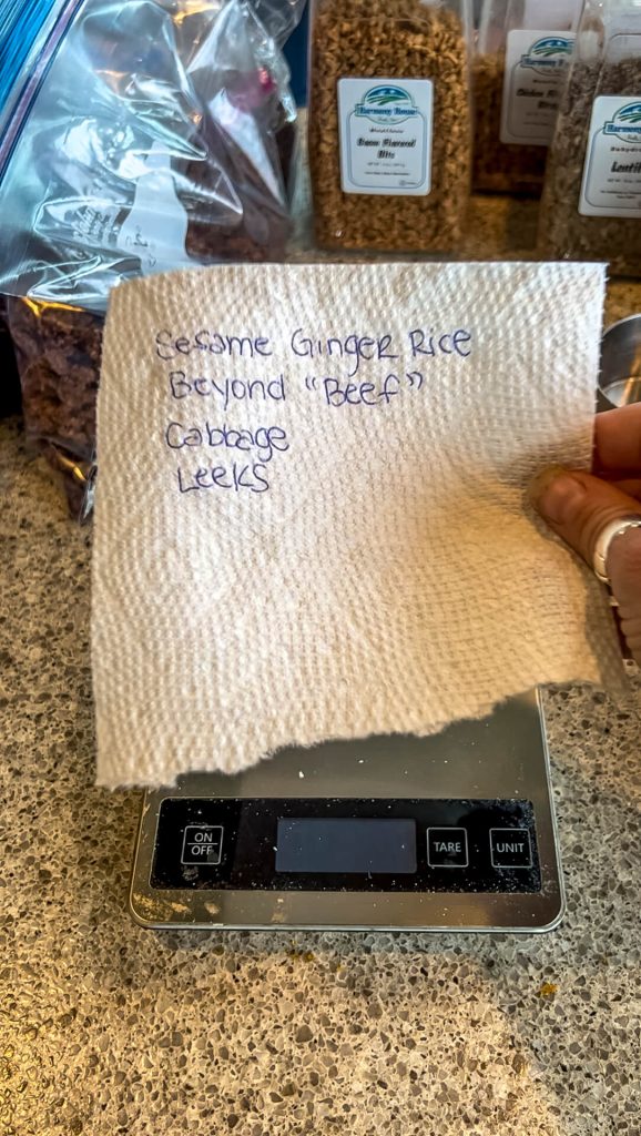 A woman's hand holding a recipe written on a paper towel over a gram scale.