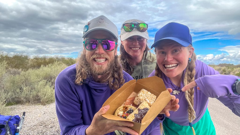 Two thru-hikers hold up a box of trail magic provided by the third human in the photo.