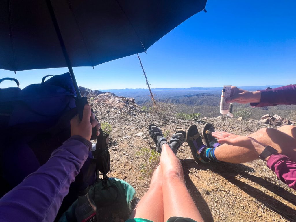 Two hikers using an umbrella for shade while they apply sunscreen in the heat of the desert.