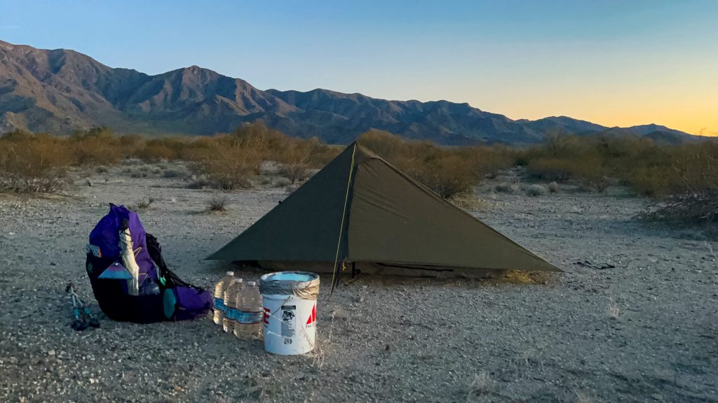 A tent, backpack, and a cache bucket in the desert.