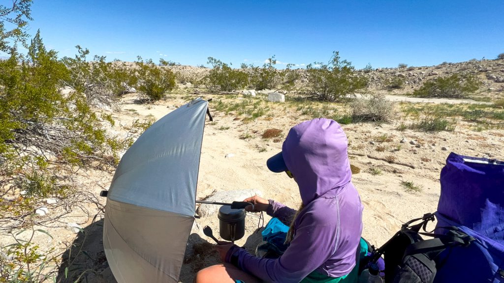 Thru-hiker using a backpacking umbrella to block the wind from her stove while she cooks.