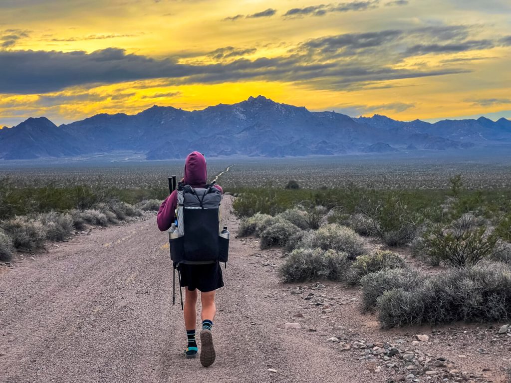 A thru-hiker making some miles through a valley on a sandy dirt road with the sunset.