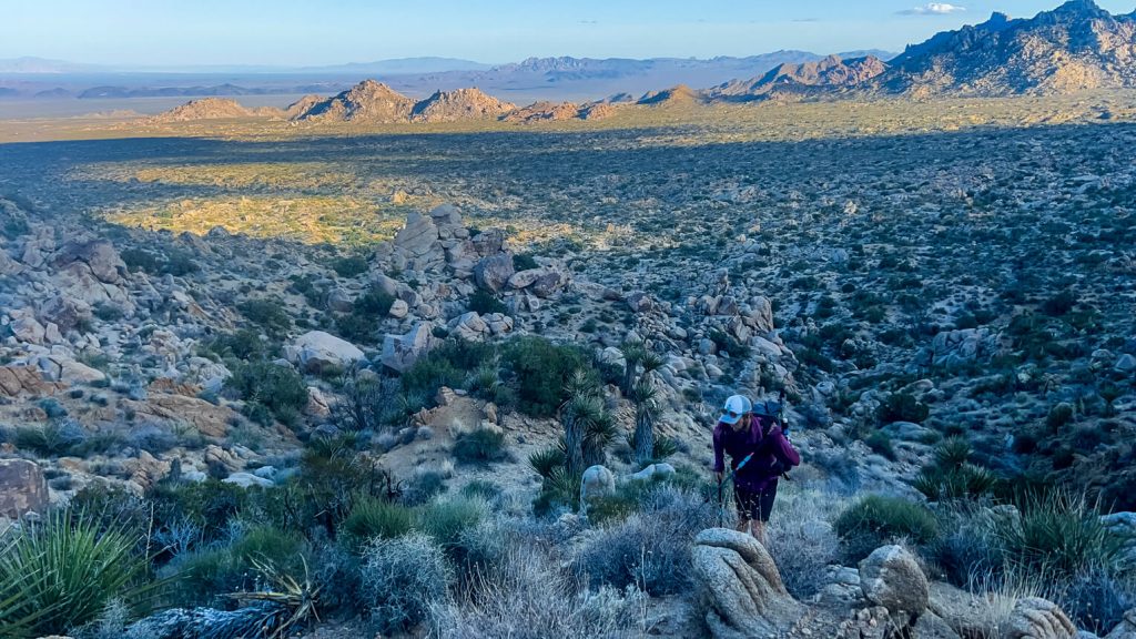 A thru-hiker climbing a low desert pass in the late afternoon shade.