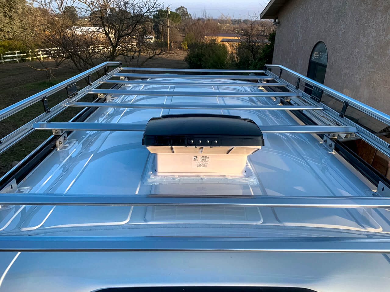 MaxxAir Fan Deluxe on the roof of a white high roof van between a roof rack.