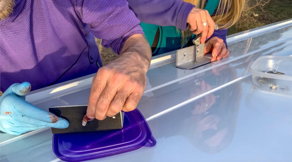 A van couple installing the Prime Design Alurack channel mounting brackets with the foam pieces, anti-seize goop (with gloves!), and an Allen Wrench.