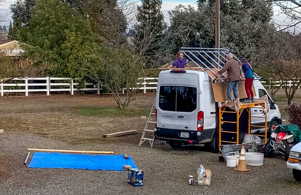 Three people working together to attach a roof rack to the top of a high roof van.
