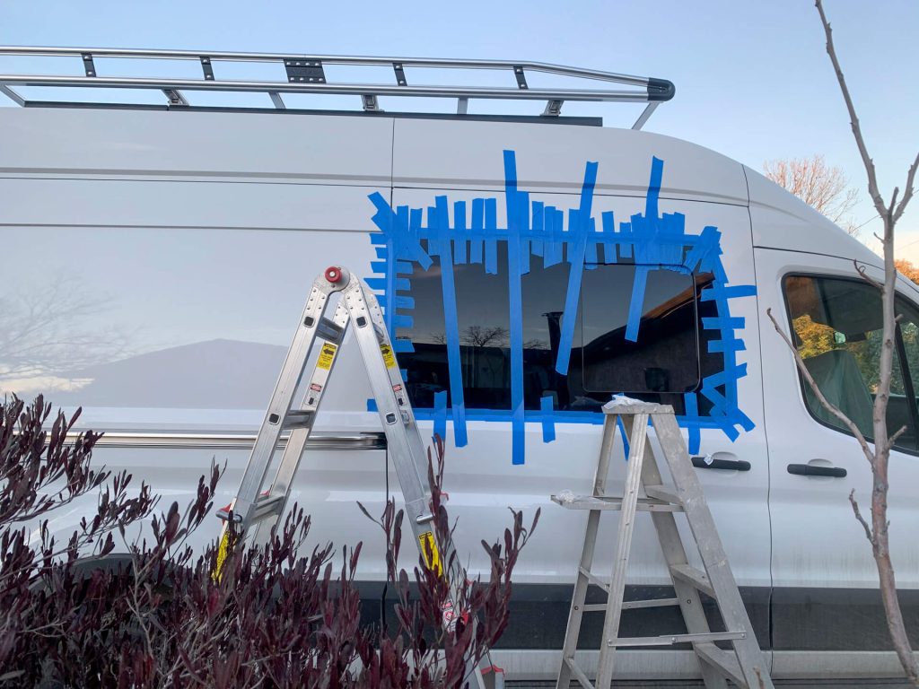Painter's Tape holding a new window in place on a Ford Transit van with two ladders in the foreground.