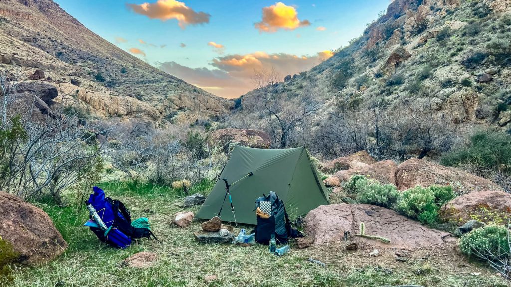 A desert campsite with a tent and two backpacks with a soft sunset.