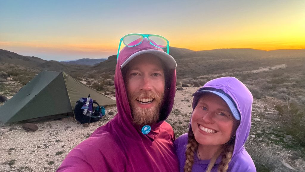 A thru-hiking couple take a selfie with their tent during sunset in the desert.