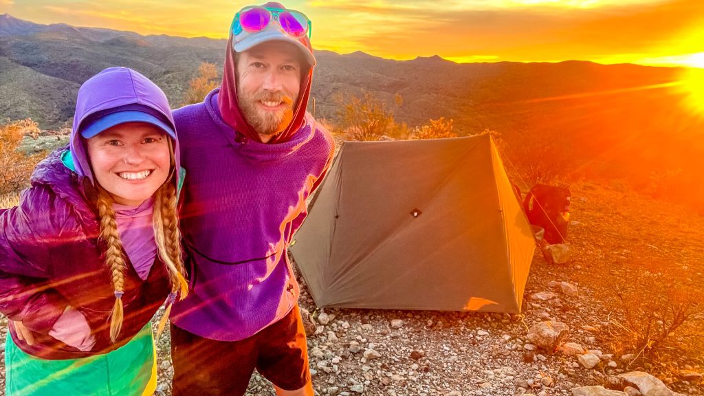 A thru-hiking couple basking in the sunset rays at their campsite with their tent in the background.