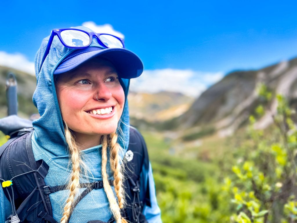 Woman thru-hiker with a wearing a backpack, sun hoodie, and sunglasses gazing off into the distance in the mountains.