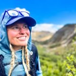 Woman thru-hiker with a wearing a backpack, sun hoodie, and sunglasses gazing off into the distance in the mountains.