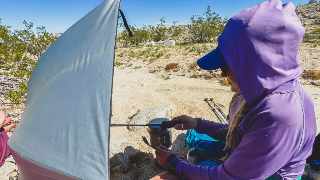 Veggie cooking lunch in the desert and using her umbrella as a wind block.