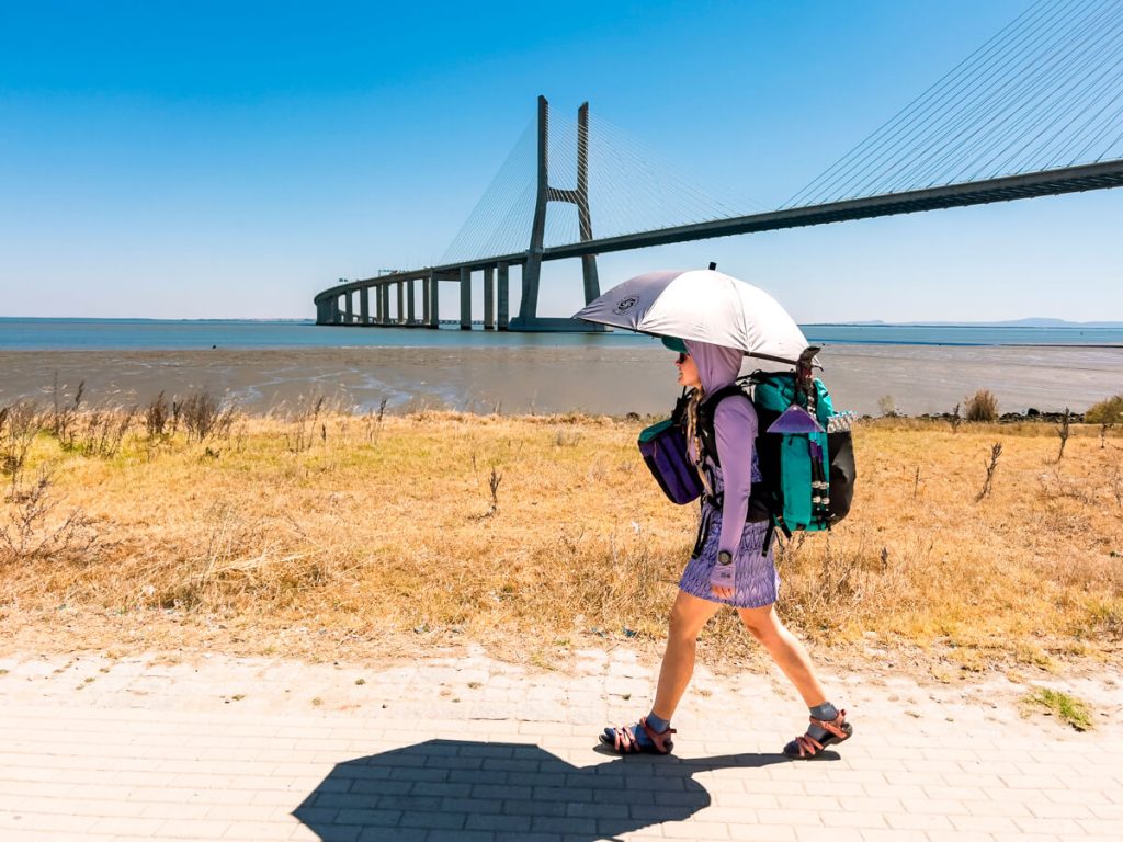 A woman camino pilgrim walks the Portuguese Camino near Lisbon in the heat with a shade umbrella.