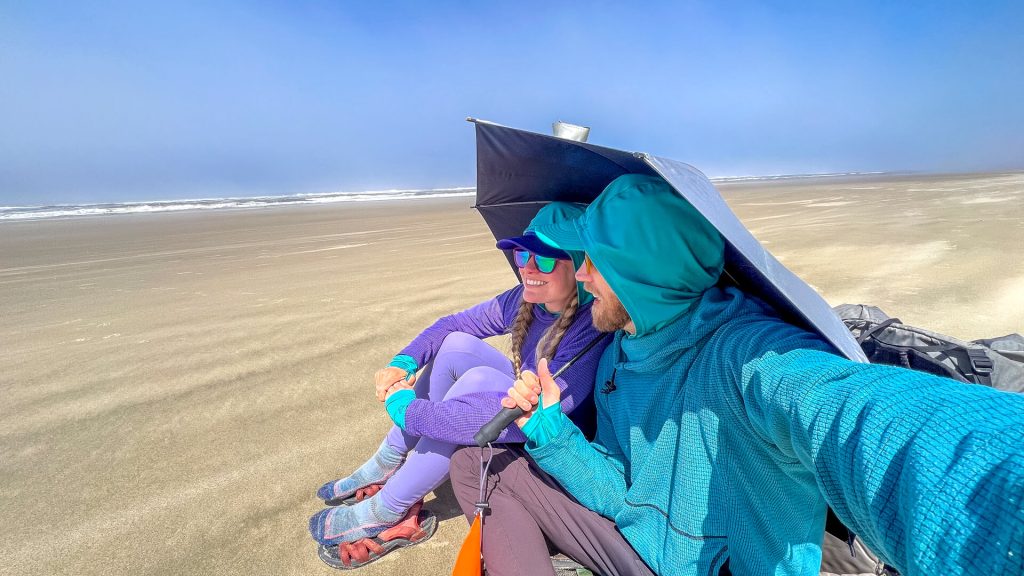 Two thru-hikers using an umbrella to block some wind on the beach for a break.